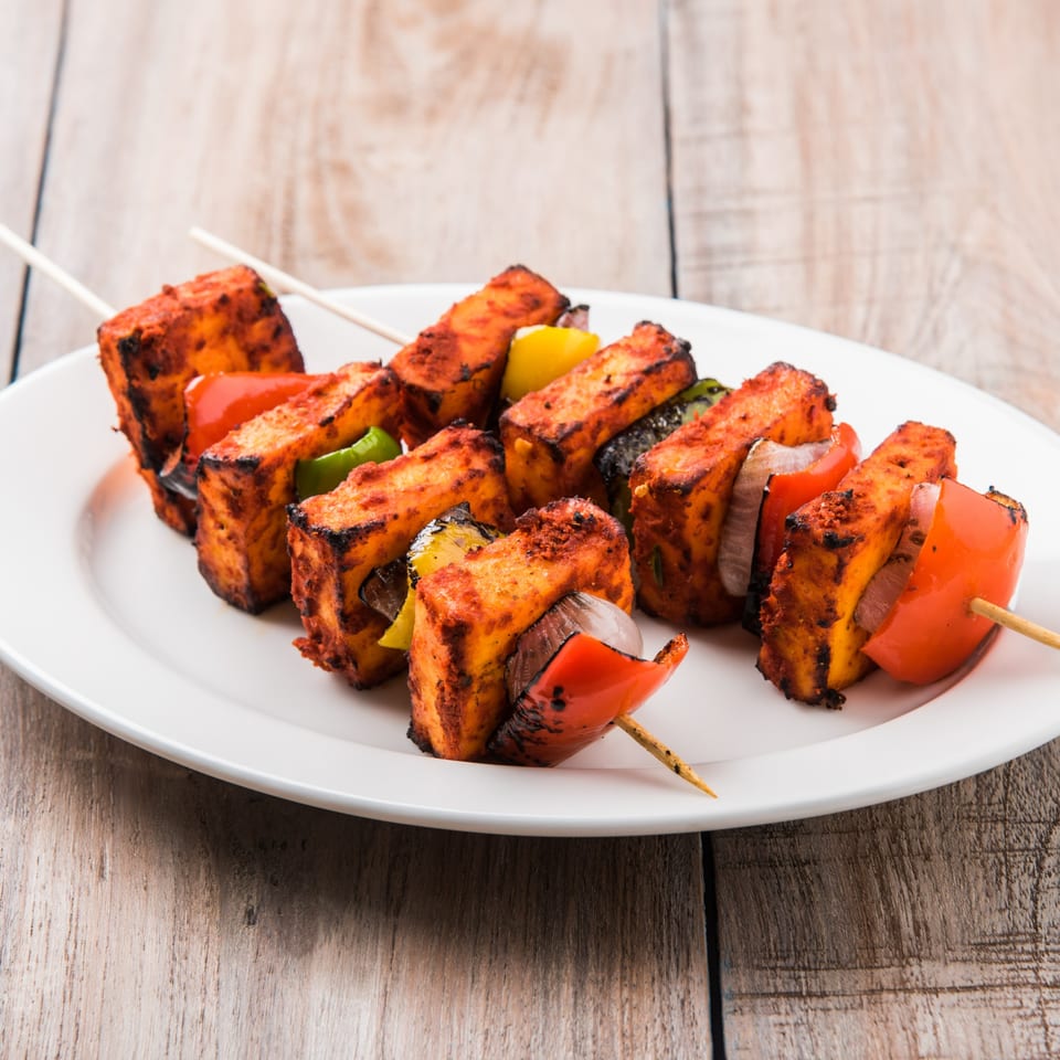Paneer tikka skewers with red and green bell peppers, onions, and cheese on a white plate.