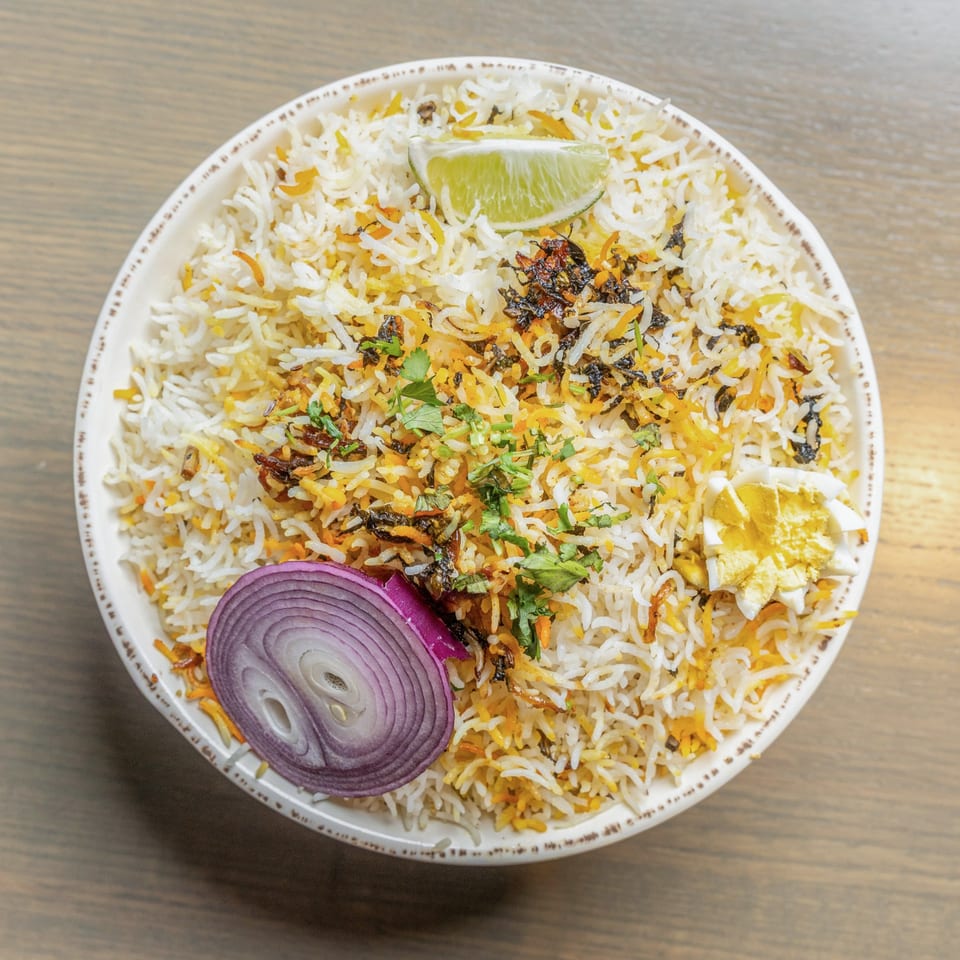 Overhead view of biryani rice with lime, red onion, cilantro, and egg in a white bowl.