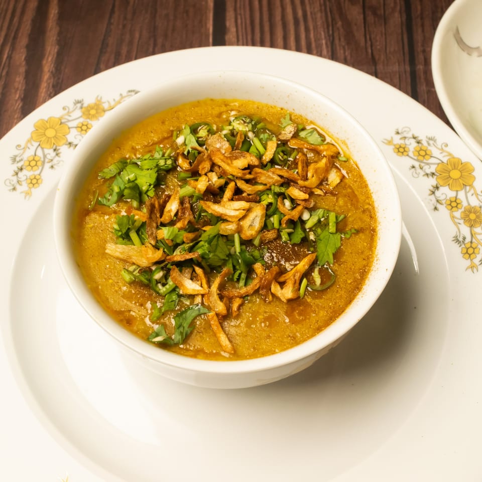 A bowl of haleem, garnished with fried onions and fresh cilantro, on a decorative plate.