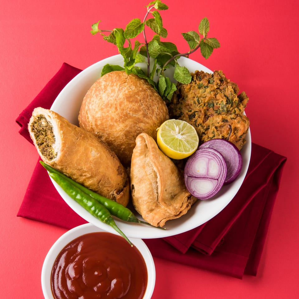 A white plate of Indian snacks including samosas, kachori, and pakora, with mint, lime, and onion.